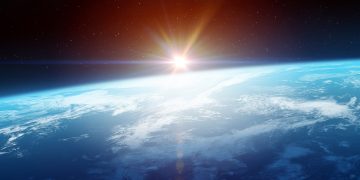 Sunrise over Earth's horizon viewed from space, with the planet's atmosphere and cloud-covered surface illuminated against a star-filled sky.